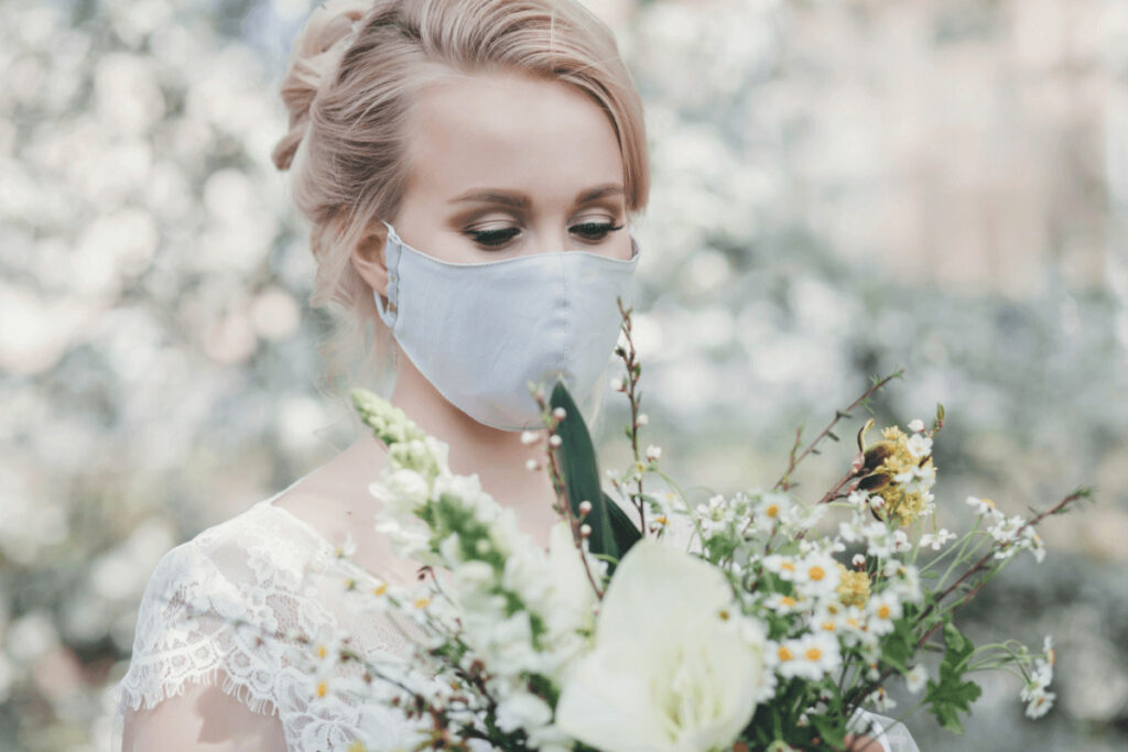 Een bruid met lichtblauw mondmasker kijkt naar het bruidsboeket dat ze draagt. 