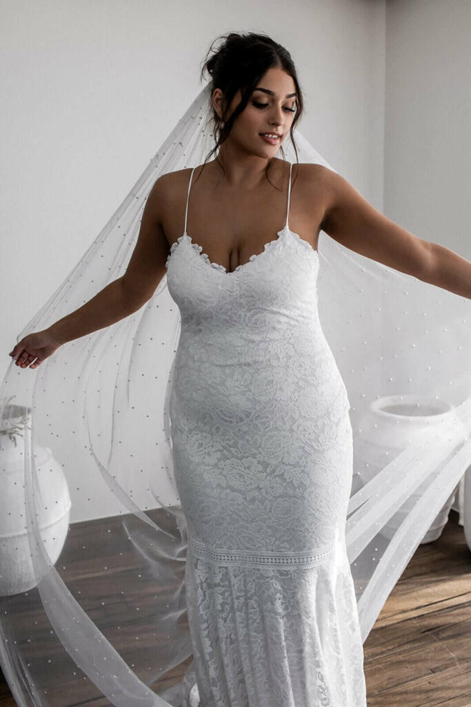 Curvy bruid draagt een kanten jurk met spaghettibandjes en een sluier bij de zoektocht van je trouwjurk.