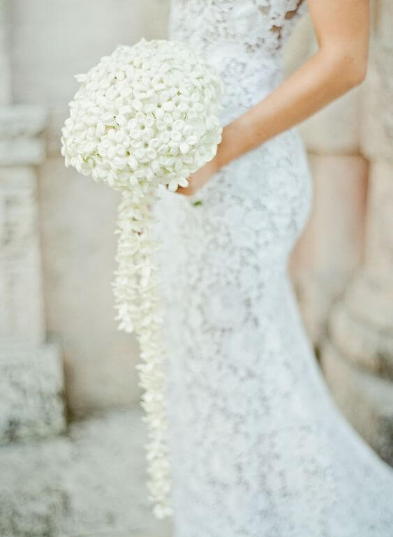 Bruid draagt een bruidsjurk in kant en een rond boeket van witte, kleine bloemen. 