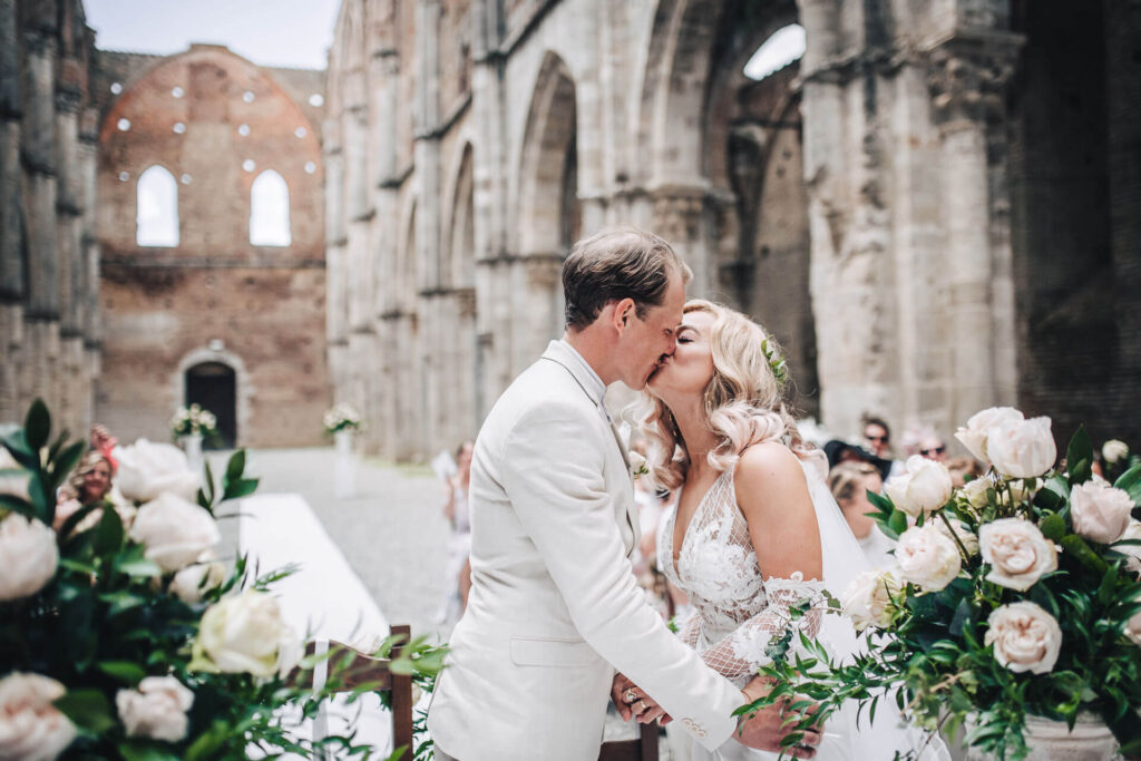 Ceremonieboekje kerkelijk huwelijk: het bruidspaar kust voor het eerst omgeven door de huwelijksgasten en prachtige witte bloemen. 


