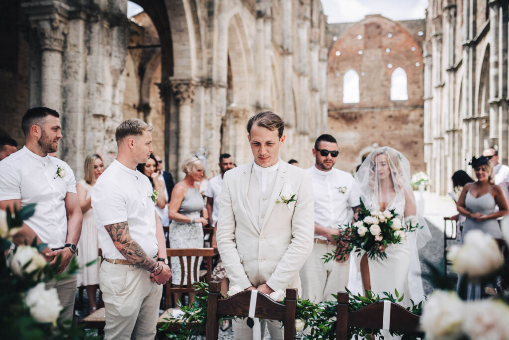 Ceremonieboekje kerkelijk huwelijk: de bruidegom in beige pak wacht, bijgestaan door zijn getuige, aan het altaar op de aankomst van de bruid. 
