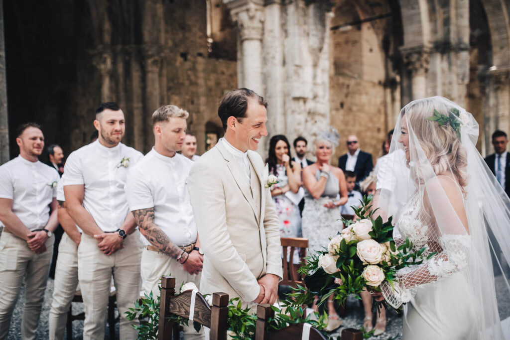 Ceremonieboekje kerkelijk huwelijk: bruidegom in beige pak begroet gesluierde bruid aan het altaar. 
