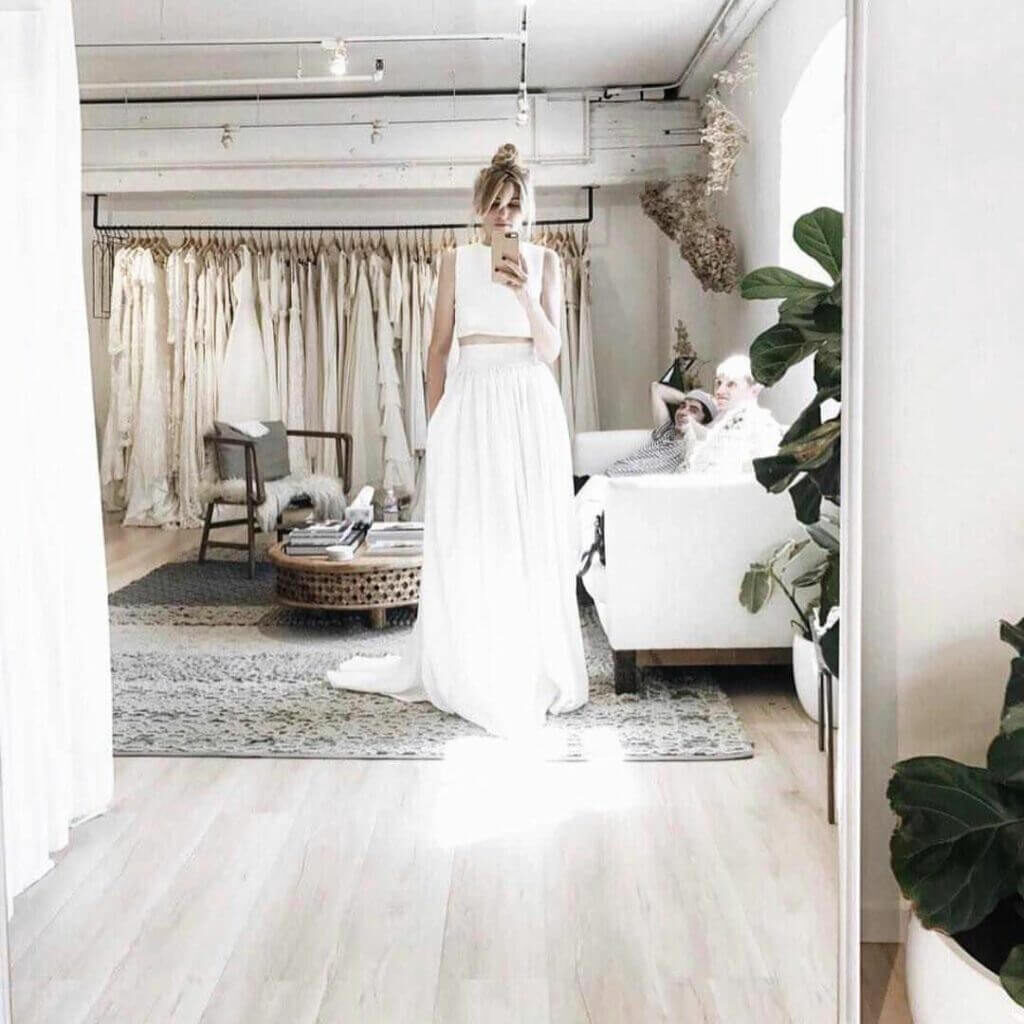 Een bruid met opgestoken kapsel maakt een selfie terwijl ze in haar tweedelige bruidsjurk voor de spiegel staat in de paskamer.