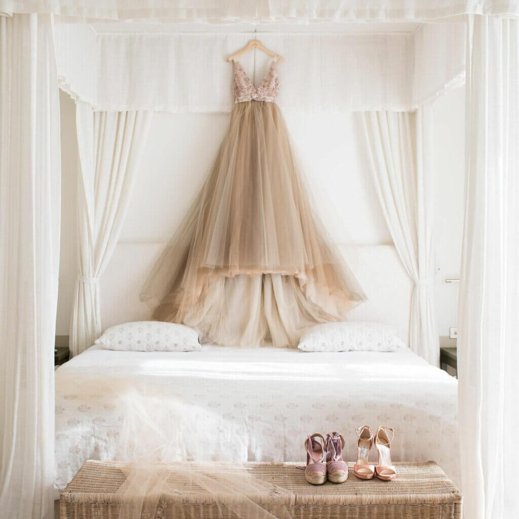 Prachtige champagnekleurige bruidsjurk hangt aan een kapstok boven het bed van de bruidssuite met twee paar bijpassende schoenen. 