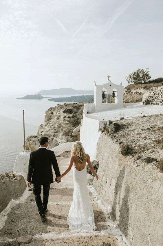 Bruid en bruidegom wandelen de trap af in het romantische Santorini en hebben zicht op een prachtig wit kerkje en de zee. 