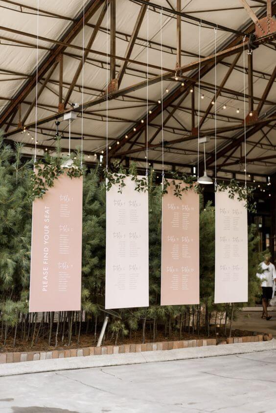Hangend seating plan op je bruiloft bestaande uit vier panelen in oudroze tinten en opgesmukt met groene takken. 