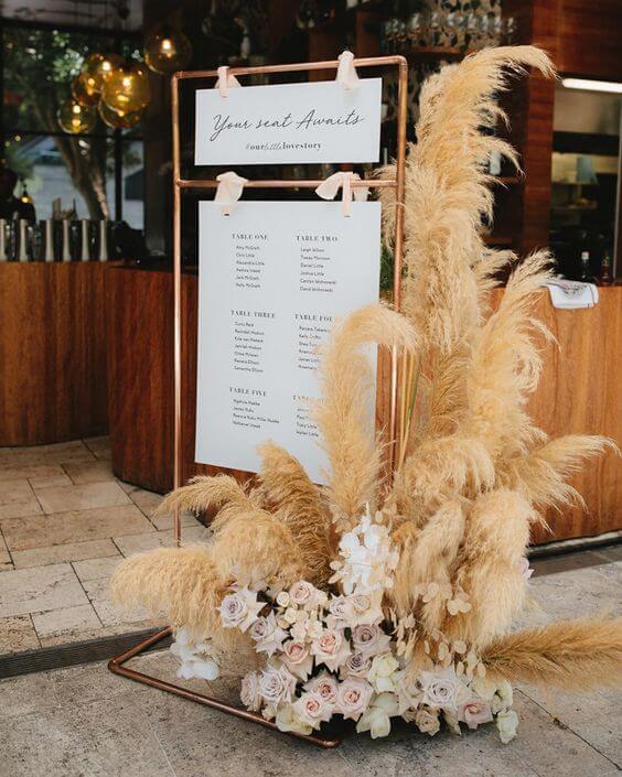 Boho-chic seating plan op je bruiloft met wit paneel en opgesmukt met een bloemstuk bestaande uit pampasgrassen en pastelkleurige rozen. 