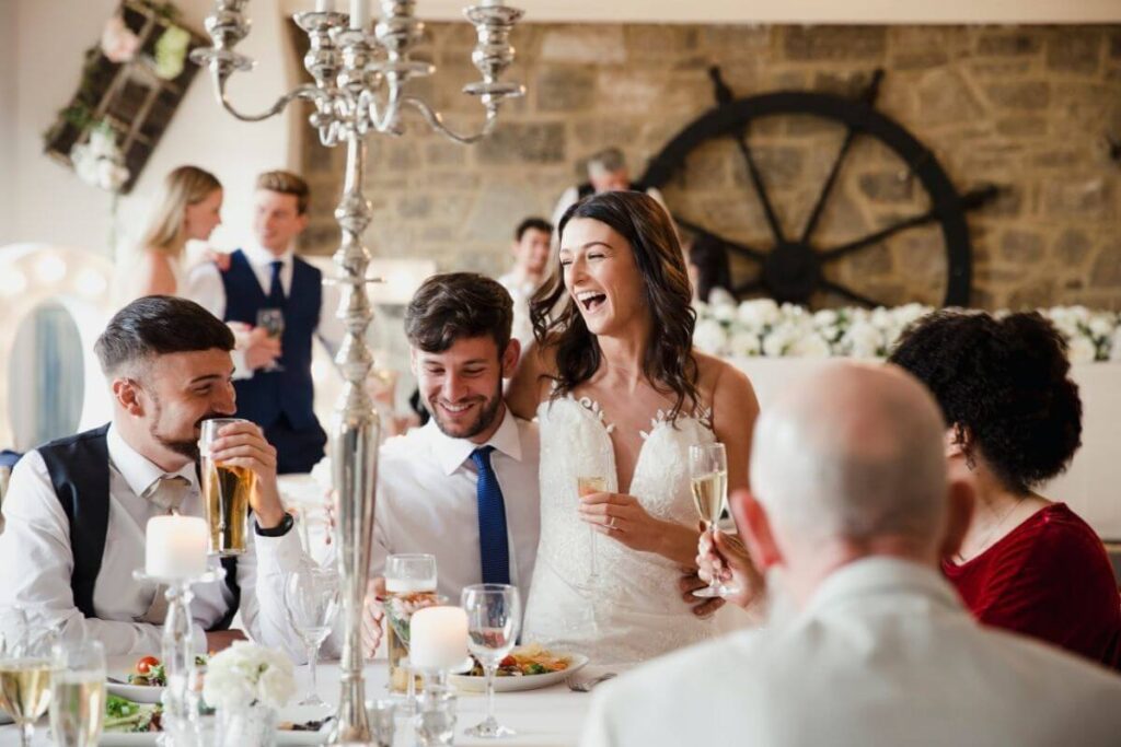 Gasten bezighouden op bruiloft: de bruid en bruidegom zitten aan een ronde tafel met hun gasten en lachen met de trouwquiz. 