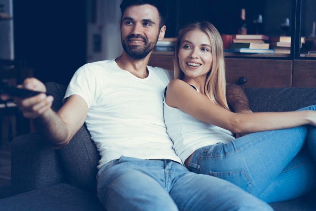Wat doen op de eerste Valentijn als pasgetrouwd stel? Koppel in wit t-shirt en jeans kijken samen op de bank naar tv. 