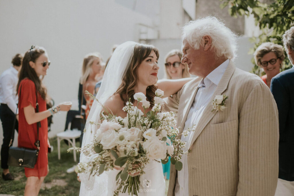 Wie nodig je uit op je bruiloft? Bruid met bruidsboeket knuffelt een oudere man met wit lang haar en beige pak. 
