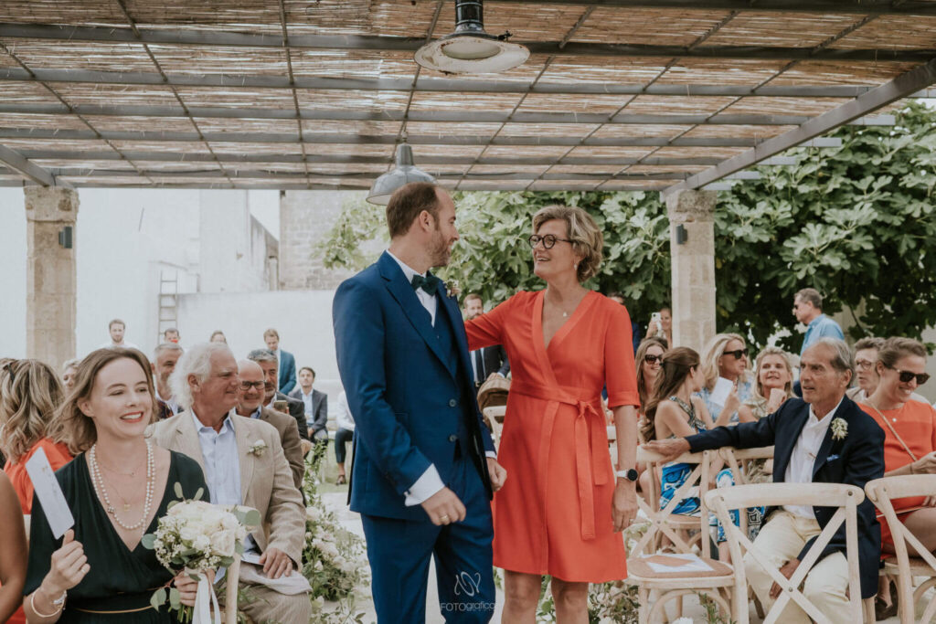 Wie nodig je uit op je bruiloft? Bruidegom in donkerblauw pak en zijn moeder in koraalrode jurk lopen naar het altaar. 