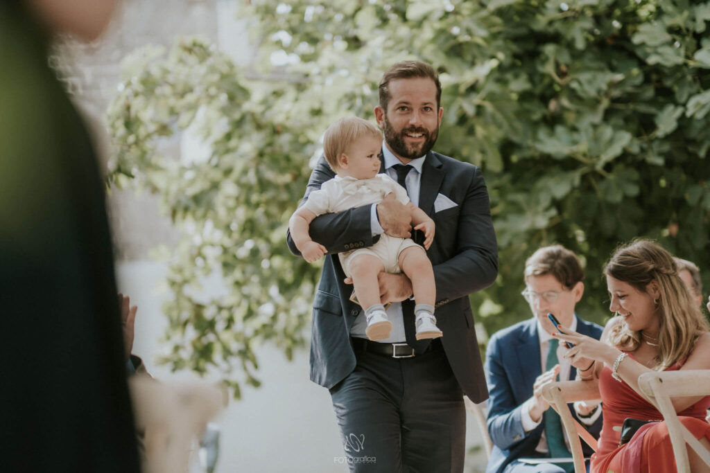 Wie nodig je uit op je bruiloft? Huwelijksgast draagt een baby in beige pakje en strikje op het gangpad naar het altaar.