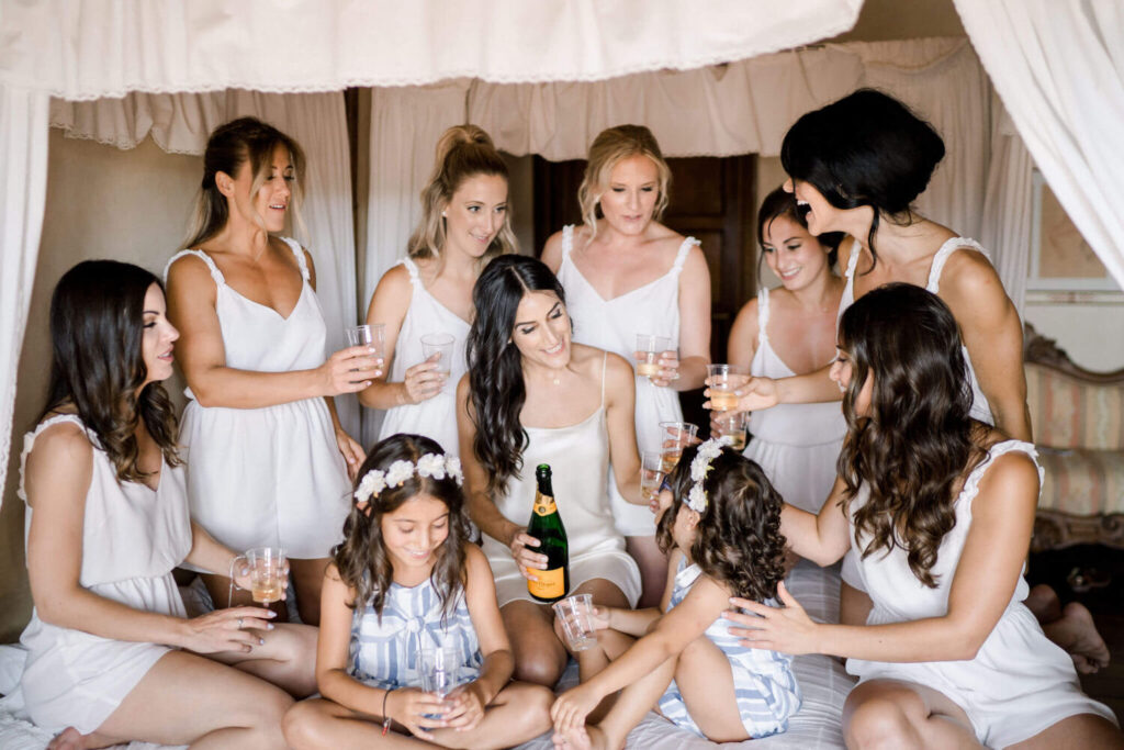 Destination wedding in Italië: de prachtige bruid zit op het bed omringd door haar bruidsmeisjes en bloemenmeisjes. Ze kraken een fles champagne.