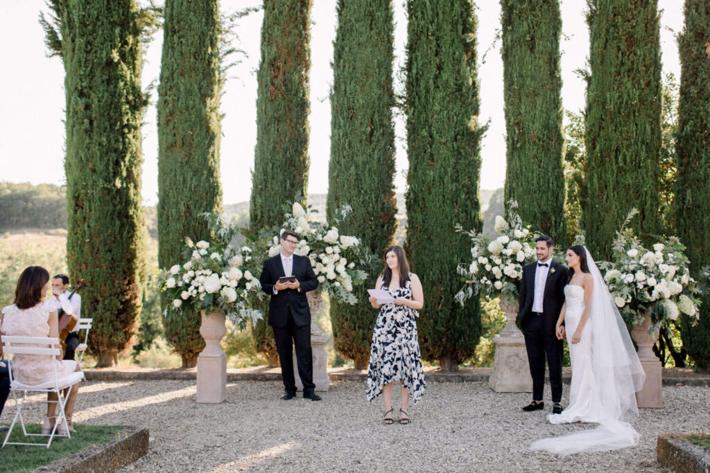 Destination wedding in Italië: een intieme huwelijksceremonie met Toscaanse cipressen op de achtergrond en prachtige witte hortensia's en witte rozen. Het bruidspaar luistert naar de ceremoniespreker die een speech voorleest. 