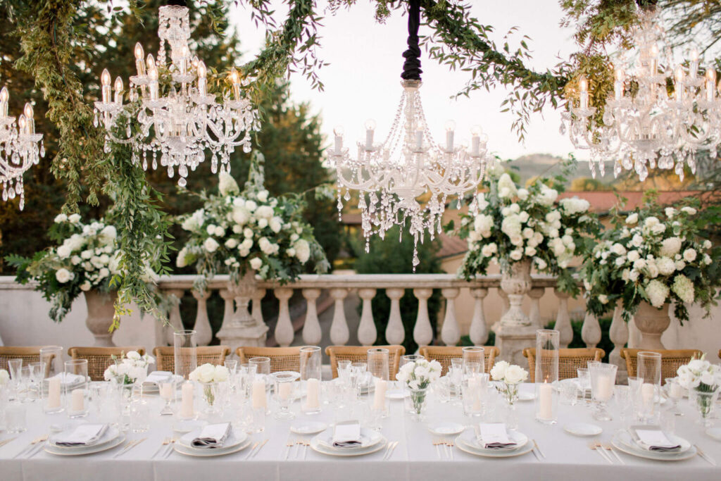 Destination wedding in Italië:  prachtige opgedekte tafel met een meterslange guirlande van groen en schitterende, kristallen kroonluchters op het pleintje voor een Toscaans landhuis.