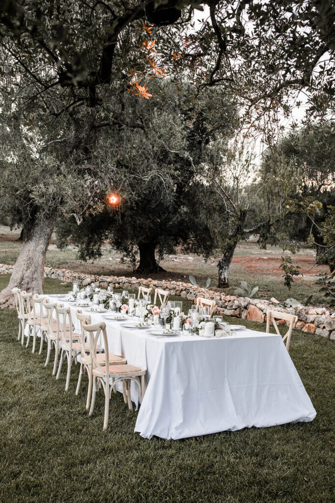 Destination wedding in Italië: een mooi opgedekte tafel in een olijfboomgaard met een lange bloemenguirlande met olijftakken en roze bloemen en witte kaarsen met op de achtergrond een robuuste olijfboom.