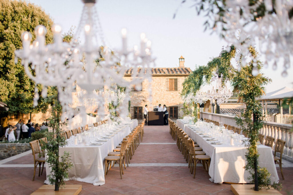 Destination wedding in Italië:  prachtige opgedekte tafels met een meterslange guirlande van groen en schitterende, kristallen kroonluchters op het pleintje voor een Toscaans landhuis.