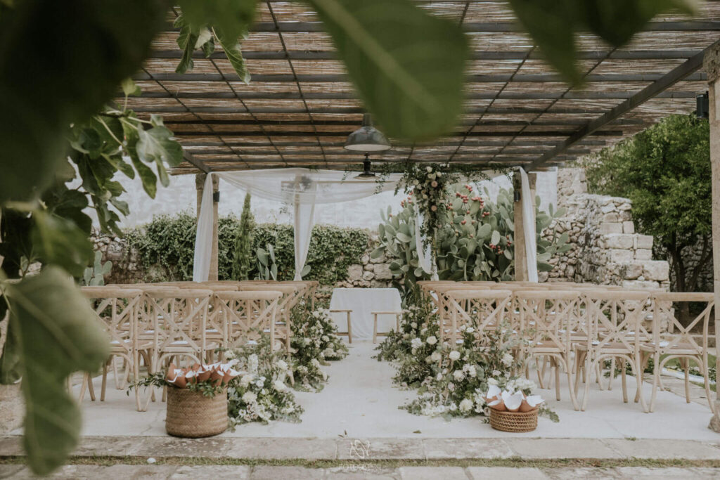 Het outdoor prieeltje voor de huwelijksceremonie is prachtig versierd met bloemstukken met groen en witte rozen. Op de voorgrond de ceremonietafel en twee houten krukjes voor het bruidspaar. Twee rotan manden met puntzakjes gevuld met witte bloemblaadjes flankeren het begin van het gangpad.