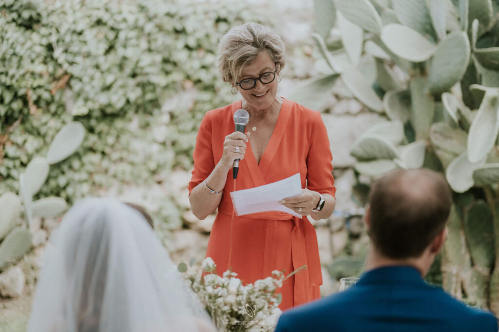 Het bruidspaar luistert naar gedichten om voor te lezen tijdens de huwelijksceremonie door de moeder van de bruidegom in een koraalrode jurk. Op de achtergrond zien we een enorme cactus als wandversiering. 
