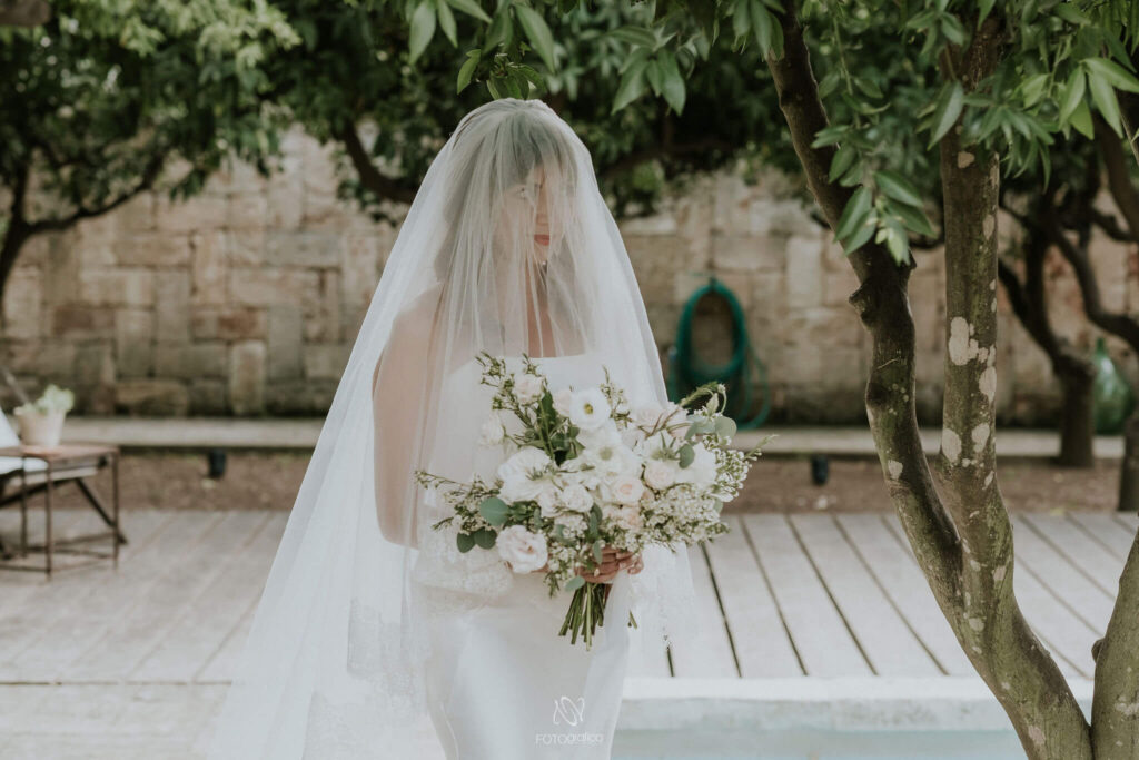 De bruid in witte jurk met sluier voor het gezicht en een boeket van groen en witte bloemen wandelt naar de outdoor ceremonieruimte voor haar huwelijk. 