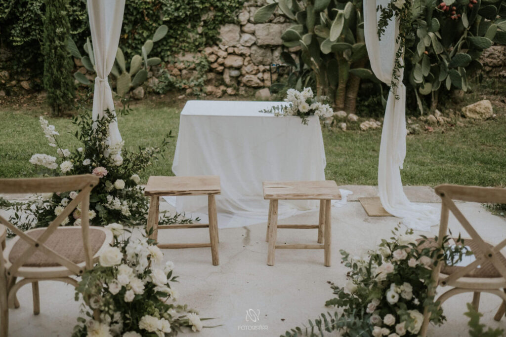 Het outdoor prieeltje voor de huwelijksceremonie is prachtig versierd met groen en witte rozen. Op de voorgrond de ceremonietafel en twee houten krukjes voor het bruidspaar.