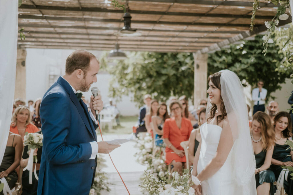 De bruidegom in donkerblauw pak spreekt met een microfoon zijn huwelijksgeloften uit. De bruid met een eenvoudige witte jurk en sluier van tule luistert en glimlacht bij het horen van zijn belofte. 