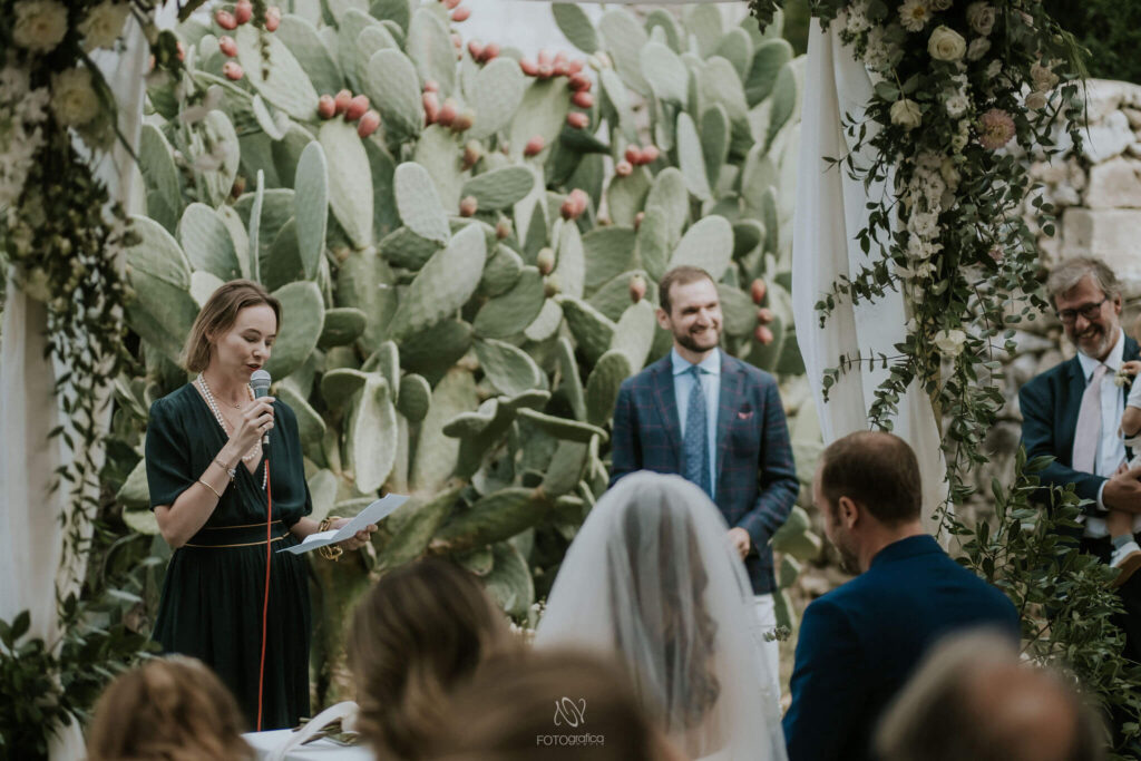 De huwelijksgasten lachen terwijl ze luisteren naar gedichten om voor te lezen tijdens de huwelijksceremonie door de zus van de bruid in een zwarte jurk. Ze zijn omgeven door eucalyptus. Op de achtergrond zien we een enorme cactus als wandversiering. 