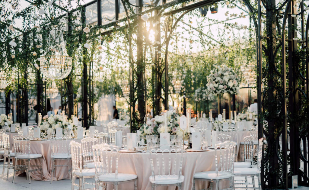 Een prachtig feestinterieur met ronde tafels in een serre met lichtroze tafellakens en met witte bloemen en kristallen kroonluchters. 