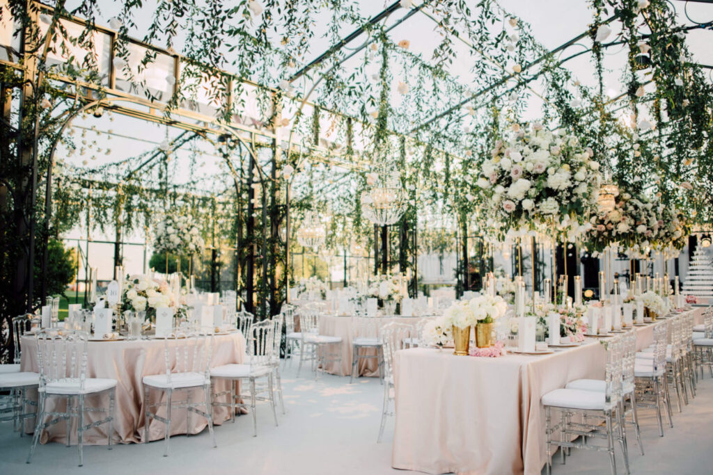 Een prachtig feestinterieur in een serre met lichtroze tafellakens en met witte bloemen en kristallen kroonluchters. 