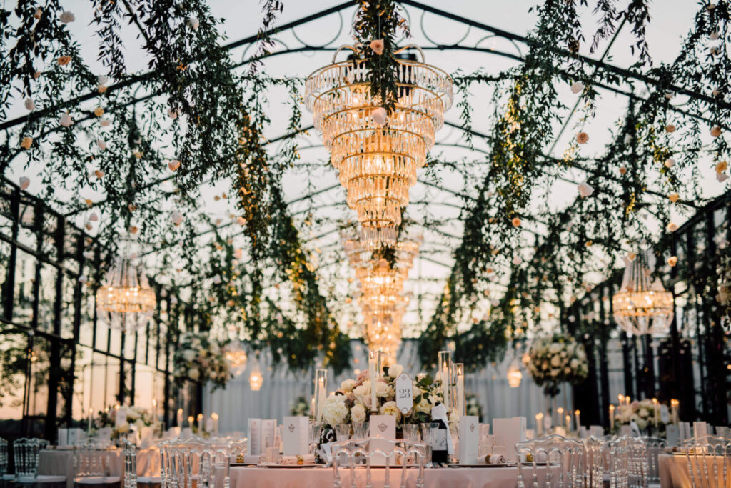 Een prachtig feestinterieur in een serre met lichtroze tafellakens en met witte bloemen en kristallen kroonluchters. 