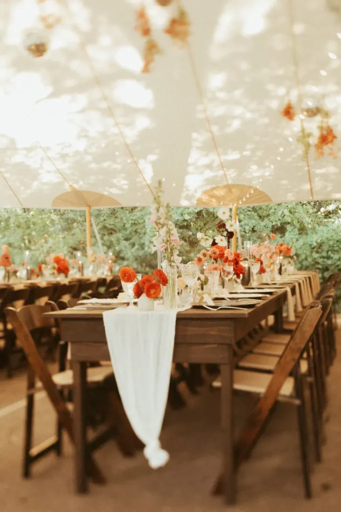 Bij zon en hitte op je trouwdag is het fijn schuilen onder een tent met houten tafels en stoelen en oranjekleurige bloemen. 