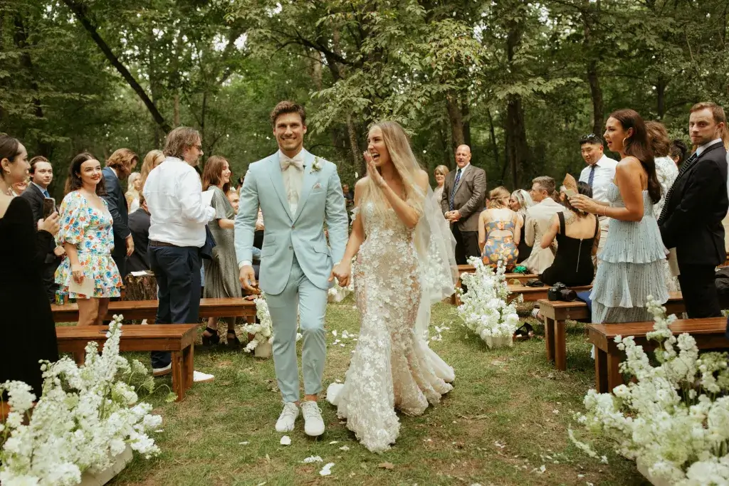 Zon en hitte op je trouwdag in het bos waarbij een blonde bruid in witte lange jurk en bruidegom in lichtblauw pak met witte sneakers zijn pas getrouwd en lopen door het gangpad in het bos. 