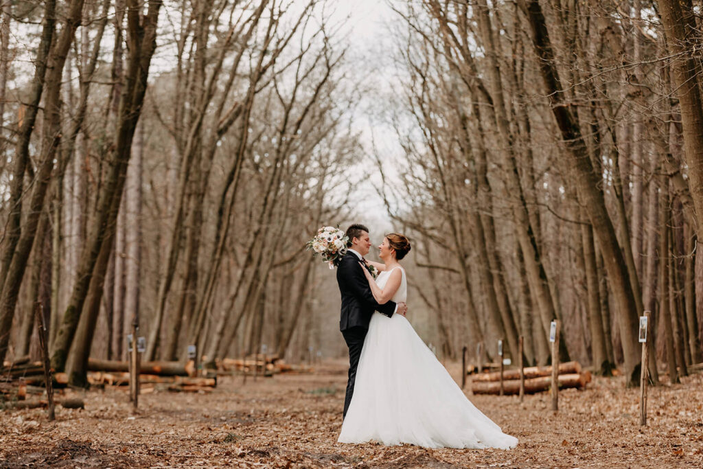 Trendy huwelijksvideo waarbij bruid in witte lange jurk en bruidegom in zwart maatpak poseren tussen de bomen in het bos. 