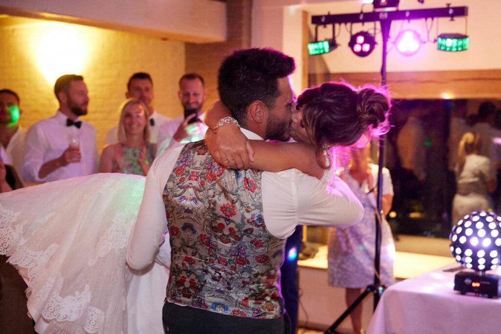 Bruidegom draagt bruid in witte trouwjurk in zijn armen terwijl ze kussen op de dansvloer op Nederlandstalige liefdesliedjes. 