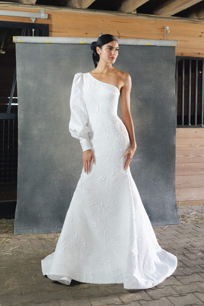 Een donkerharige bruid draagt een witte trouwjurk met een asymetrische halslijn en een ontblote schouder. 