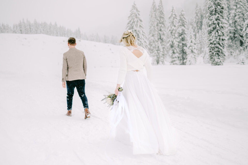 Blonde bride in white wedding dress and groom walking in the snow surrounded by snow-capped trees. 