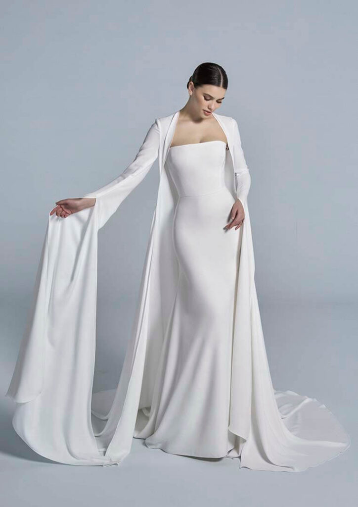 Een donkerharige bruid draagt een eenvoudige witte trouwjurk en een lange cape met lange mouwen met een split. 