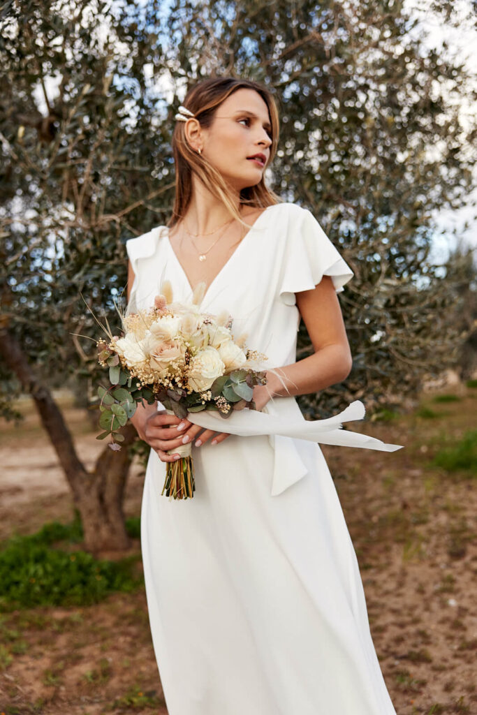 Een blonde bruid draagt een witte lange  trouwjurk met kapmouwtjes uit de bruidscollectie LolaLiza en Flair  en draagt een boeket met droogbloemen.