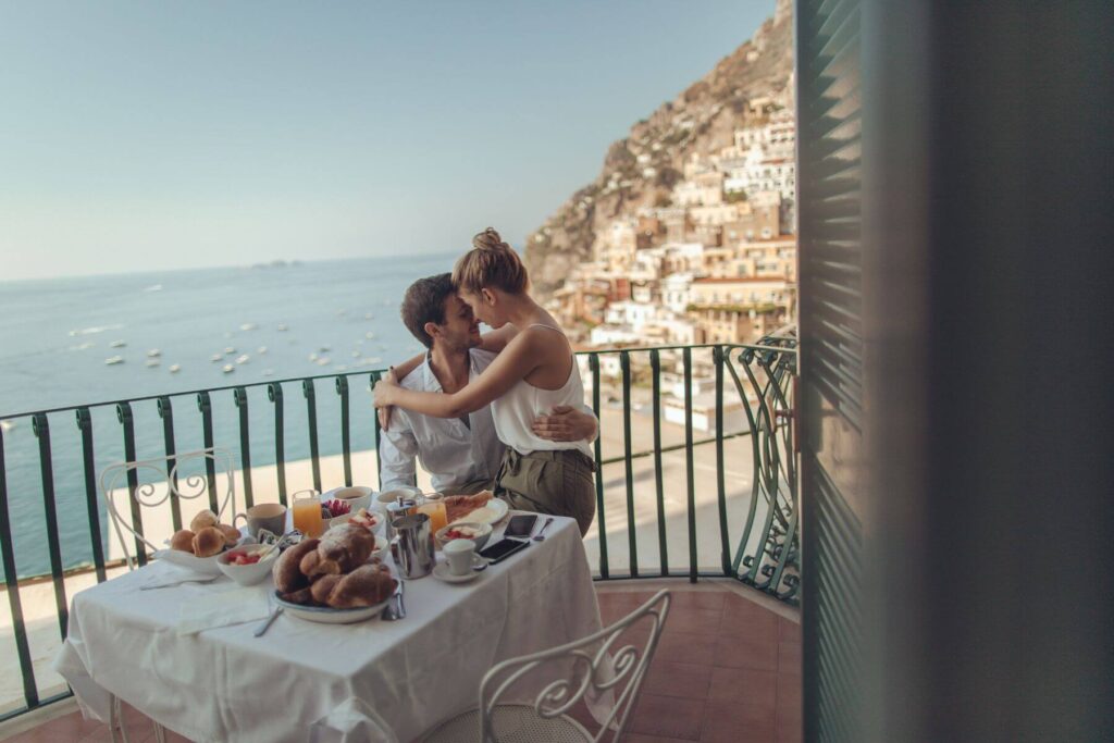 De ideale huwelijksreis waarbij een kersvers bruidspaar ontbijt op een terras met een prachtig zicht op een baai aan zee. 