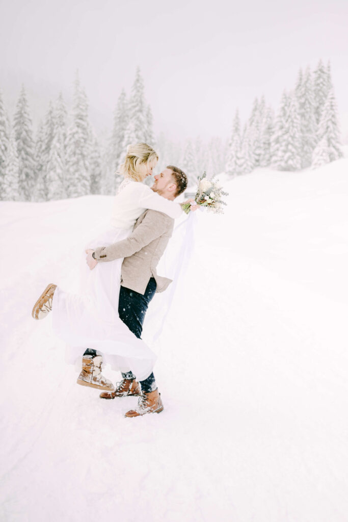 Blonde bride in white wedding dress and red head groom lifting up his bride surrounded by snow-capped trees in the mountains. 