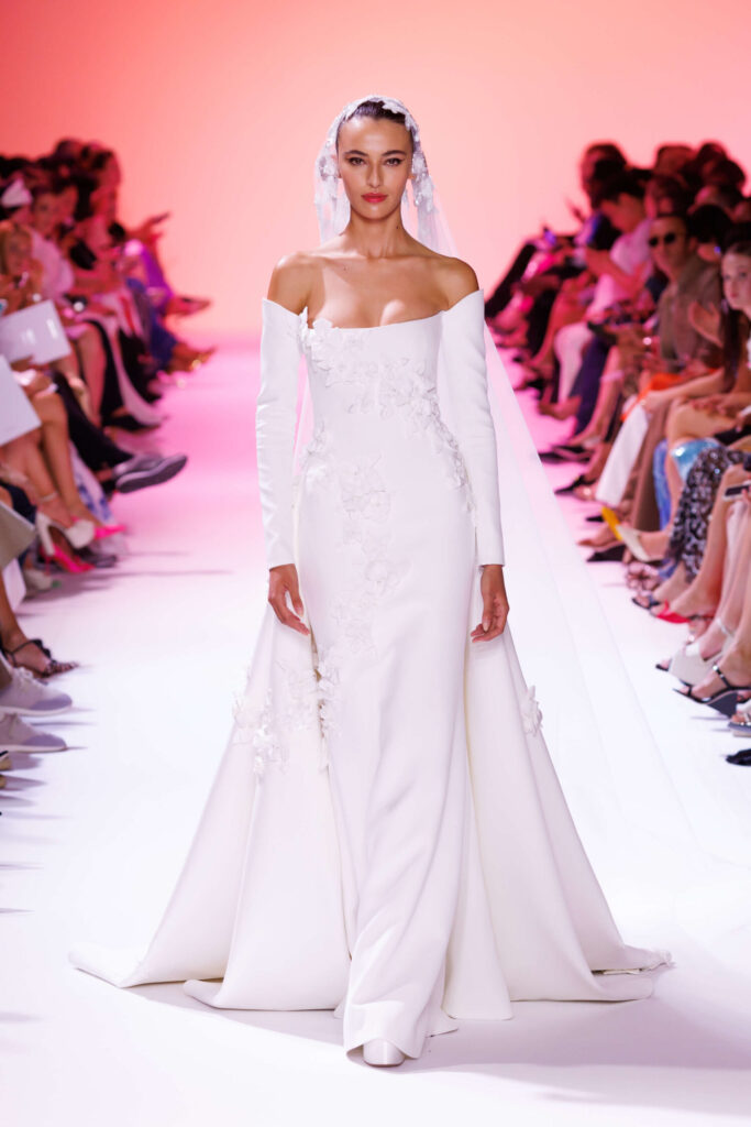 De grootste trends van Paris Haute Couture Week waarbij een model een witte jurk met bootcut hals en een sleep draagt. 